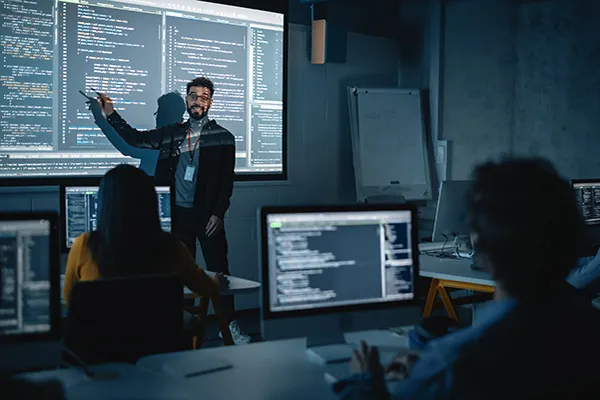 Teacher Giving Computer Science Lecture to Diverse Multiethnic Group of Female and Male Students in Dark College Room. Projecting Slideshow with Programming Code. Explaining Information Technology.