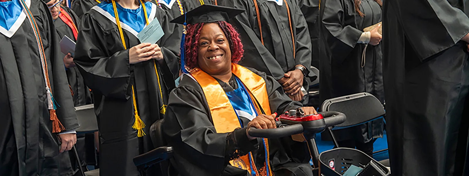 Female student in commencement attire, seated in motorized wheelchair, smiles among her fellow graduates.