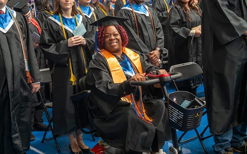 Female student in commencement attire, seated in motorized wheelchair, smiles among her fellow graduates.