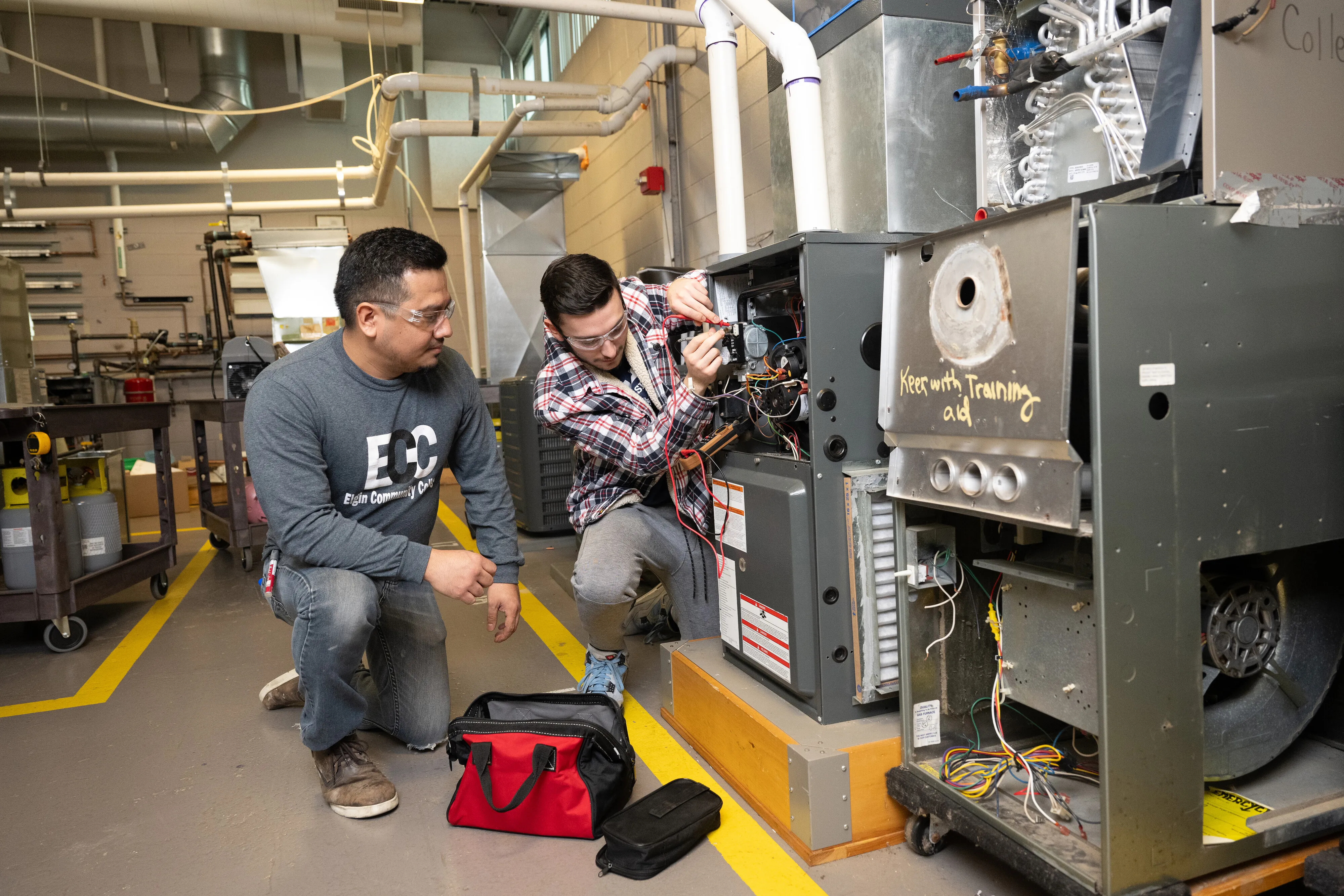 Two individuals work on an HVAC system in a workshop.
