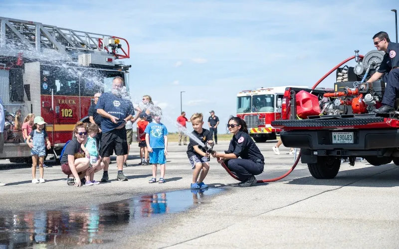 Firefighters assist children as they spray a firehose at a past 
