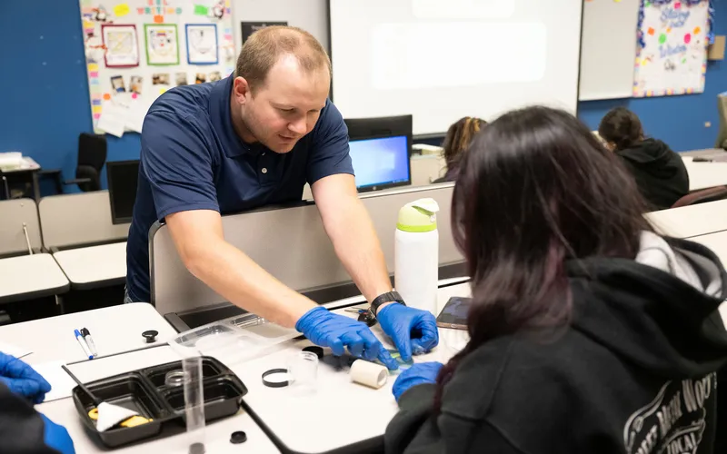A man wearing a blue shirt and blue gloves is helping a student sort through small objects from a black kit.