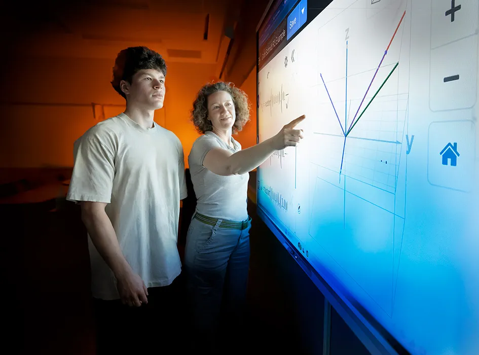 Teacher and student interact with a large digital display, examining a graph with multiple lines in a classroom setting.