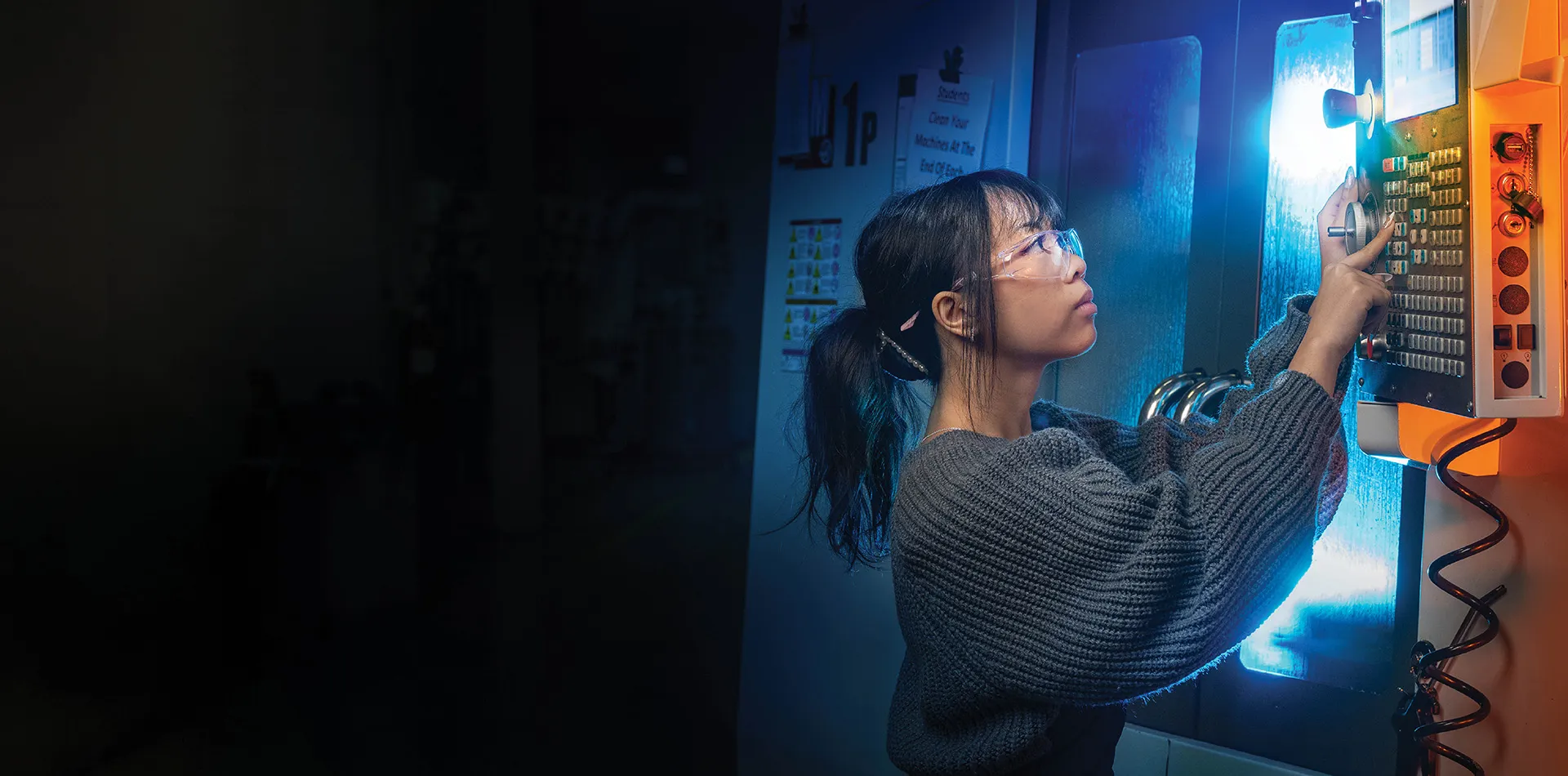 Person wearing safety glasses operates a control panel on an industrial machine, illuminated by blue and orange lighting in a dim workspace.