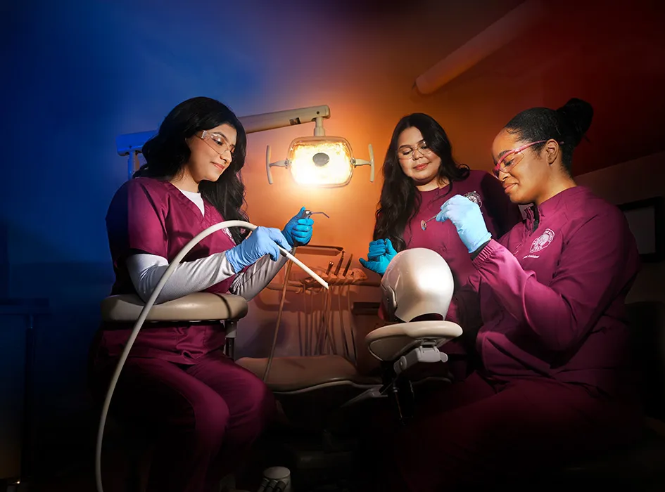 Three dental students in scrubs practice procedures on a mannequin head using dental tools in a clinical lab.
