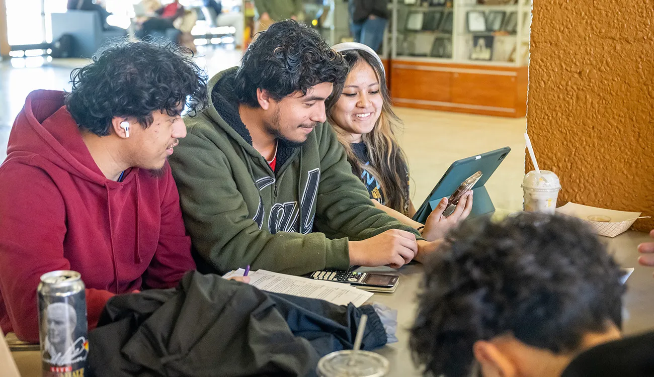 Student smiles while working on a desktop computer in a classroom lab, with classmates using computers in the background.