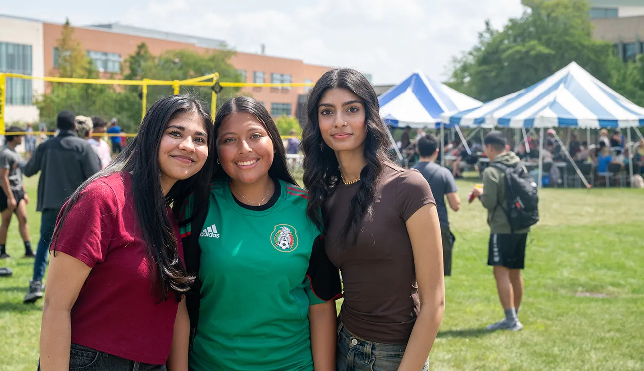 Three students smile together on a campus lawn during an outdoor event, with tents and a volleyball net in the background.