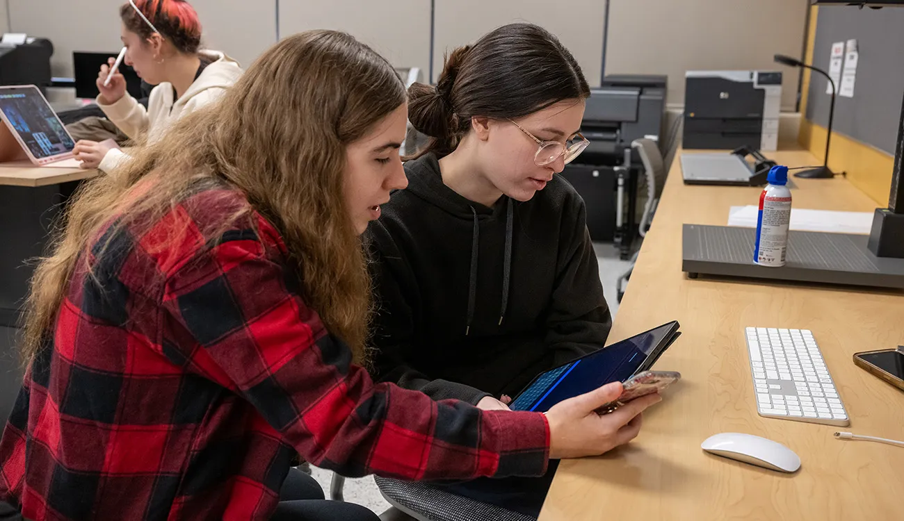 Two students collaborate at a desk, reviewing content on a tablet while working in a computer lab.