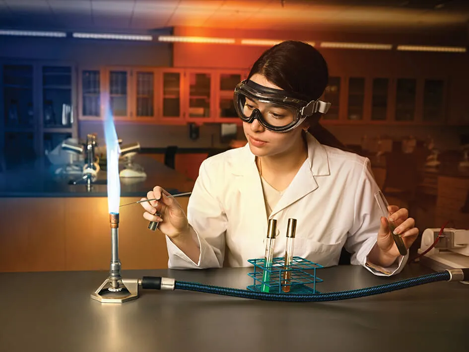 A student wearing goggles and sitting at a table using a bunsen burner