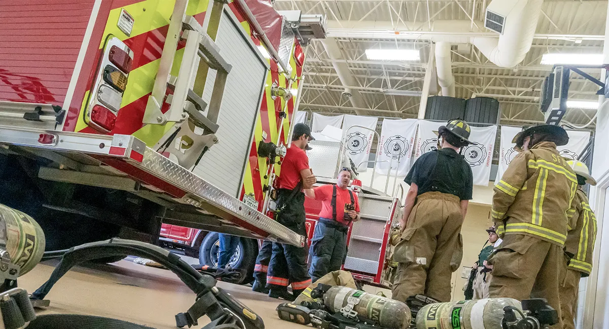 Group of students in partial gear standing next to a fire engine inside an apparatus bay