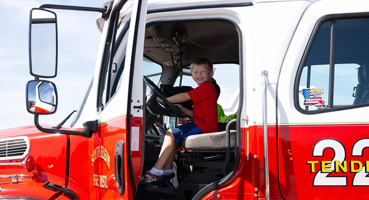A Child sitting in the drivers set of a hook and ladder fire truck at a Touch a Truck event.