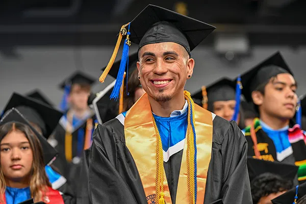 Un graduado sonriente, ataviado con toga y birrete negros y vestimenta académica azul y dorada, se encuentra entre sus compañeros graduados.