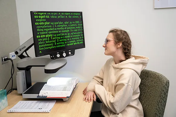 Student using a desktop video magnifier to enlarge and read text from an open book at an accessibility workstation.