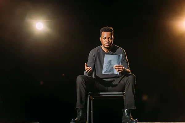 Person on a stage sitting on a chair and reading a script