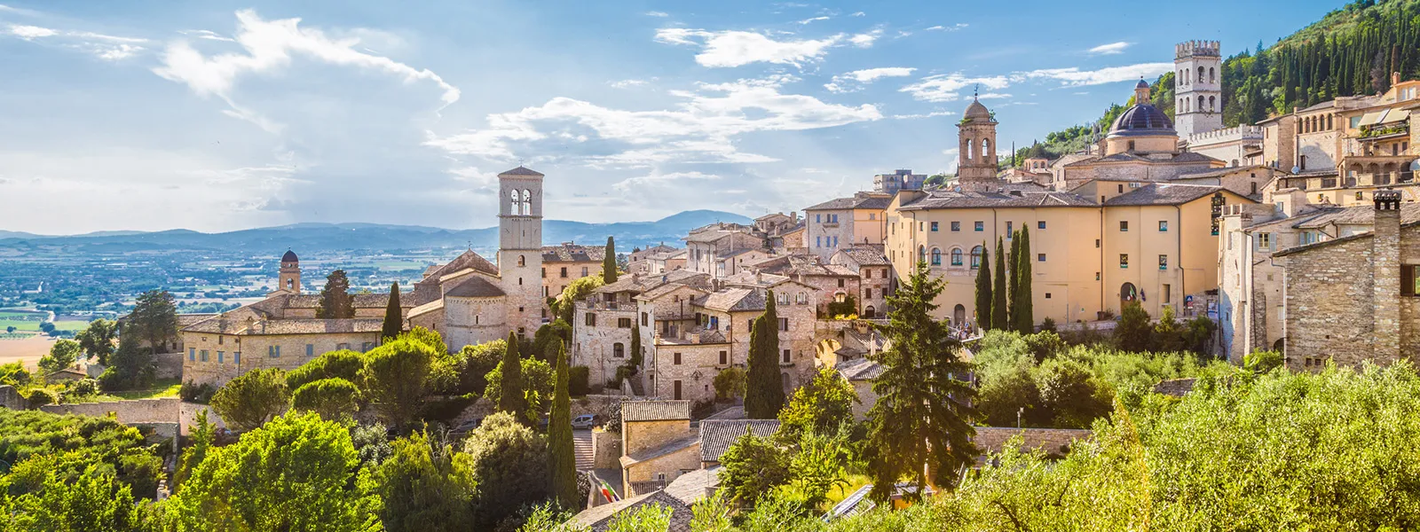 Panoramic view of a historic Italian hill town with stone buildings, church towers, cypress trees, and a green valley beneath a bright blue sky.
