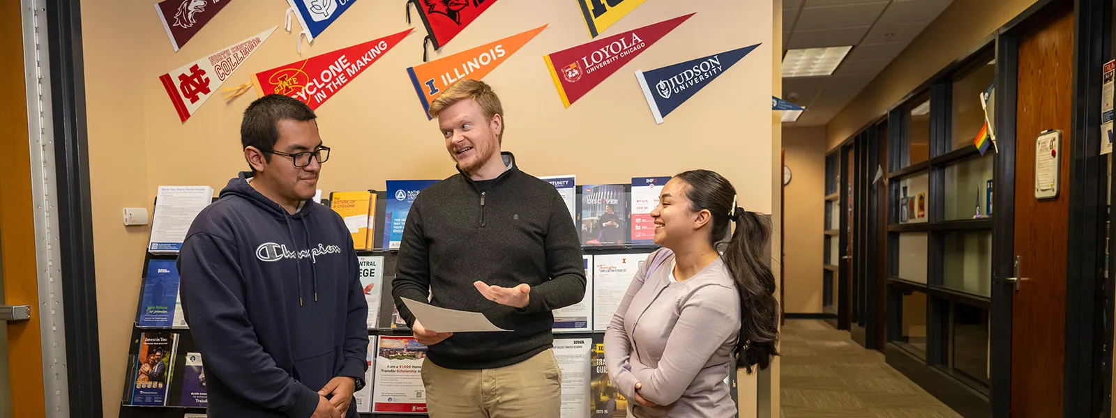 Three students stand in a college transfer center, smiling and talking beneath university pennants and beside a display of transfer resources.