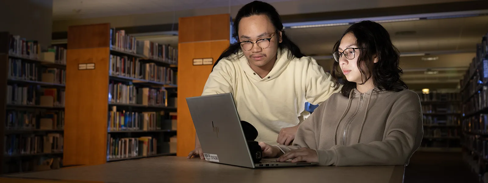Two students work on a computer in a dimly lit library