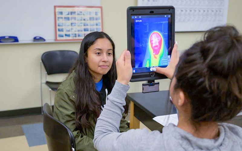 A female student uses an infrared thermal scanner on another female student seated in a classroom.