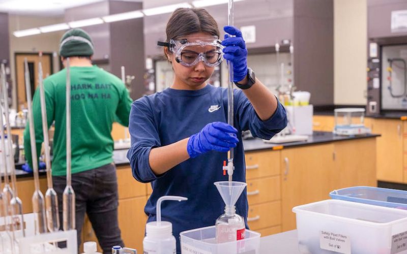 Una estudiante, con gafas de seguridad, está mezclando productos químicos en un frasco de vidrio en el aula.