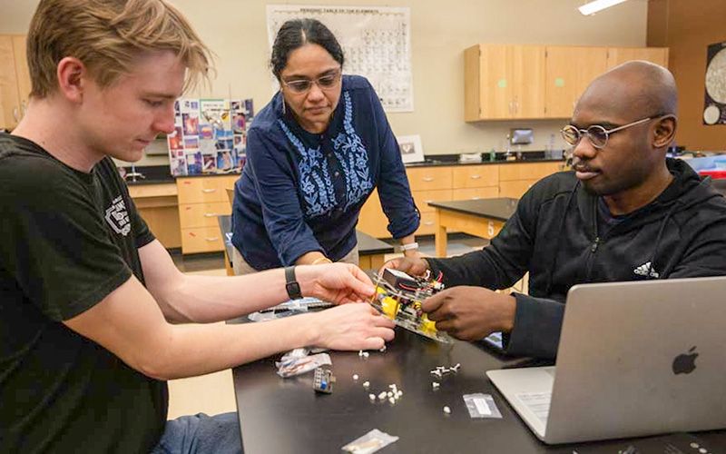 A female instructor engages with two male students as they examine electrical components at a classroom table.