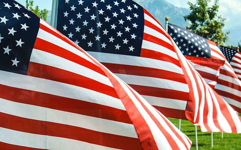Several American flags displayed outdoors with trees and mountains in the background.